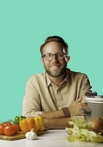 Photo of Matthew Bounds in front of a sea green background, arms crossed on a table, with peppers, tomatoes, onions, lettuce and a crockpot.